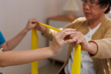 Caregiver supporting senior woman performing arm rehabilitation exercises with a yellow resistance band for physical therapy at home