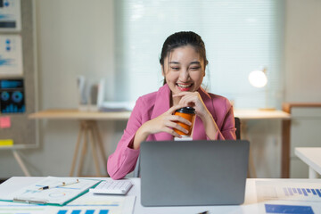 Asian businesswoman laughing during remote online video call using laptop, holding coffee cup at modern office workstation