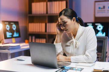 Stressed businesswoman working on laptop at desk, experiencing headache or mental exhaustion during late night overtime