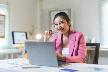 Obraz premium Young Asian professional woman in a pink blazer smiling confidently while using a laptop at her desk, working efficiently