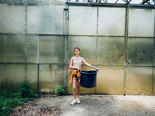 Fototapeta premium Woman in front of greenhouse holding a large black bucket, casual clothing and sneakers, gardening scene with plants and soil, outdoor agriculture and horticulture activity.