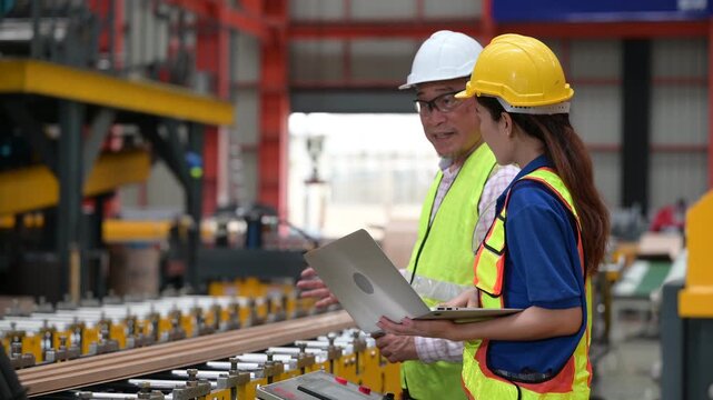 Senior Asian male supervisor pointing to train female worker on machinery in metal sheet factory. Industrial engineering mentorship, safety gear and manufacturing teamwork concept.