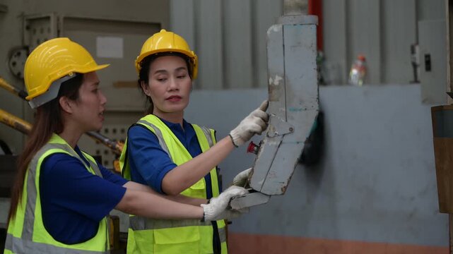 Two Asian female factory workers measuring metal sheet with tape measure. Team of engineers checking dimensions and recording quality control data in industrial manufacturing plant.