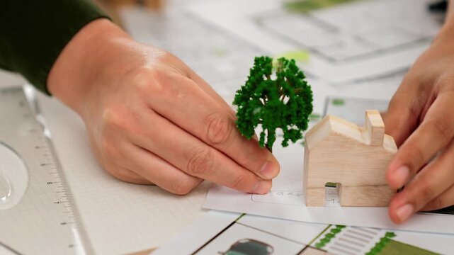 A video of architect using tree and home model to measure blueprint. Architect designing house construction on a table at studio, with architectural plan scattered around. Focus on hand. Delineation.