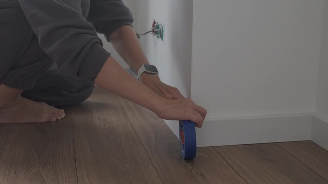 Person applying masking tape along baseboard on wooden floor indoors. Concept of interior renovation, wall painting preparation, home repair and DIY finishing work.