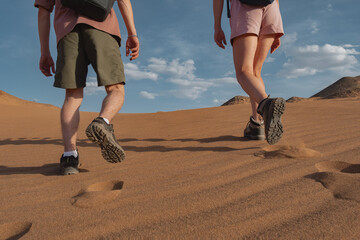 A couple in hiking shorts and boots ascend a sandy slope in the Mongolian desert. Low rear-angle shot focuses on boot soles, footprints, and their shadows on the warm sand during the climb.