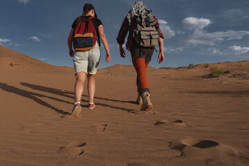 A man and woman in bright gear hike up a sandy hill in the Mongolian desert during golden hour. Rear-view shot captures their long shadows on the sand under a vast blue sky with white clouds.
