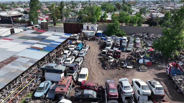Forward drone aerial of junkyard with rows of scrapped cars, warehouse structure, and residential neighborhood with trees backdrop