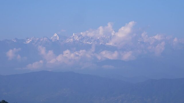 Scenic view of faded green hills and shiny snowy white mountains with clouds and a plane flying by.