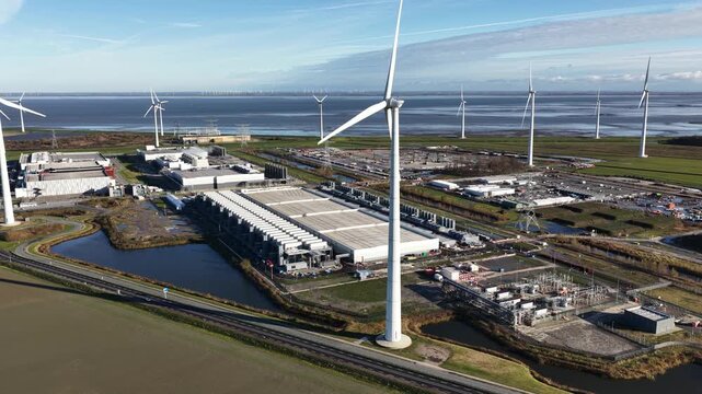 Aerial drone video of wind turbines before a large energy consuming data center in Eemshaven, showing the relationship between renewable power generation and modern computing infrastructure.