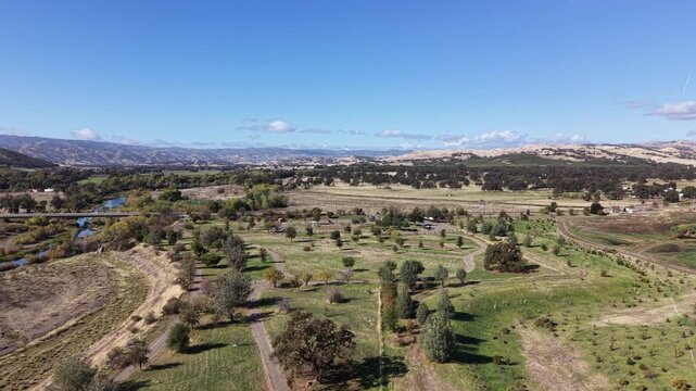 Drone performs a gentle right-to-left pan above winding dirt paths, open fields and Cache Creek, with distant hills under a clear sky in Capay, California.