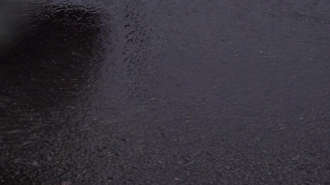 Water spray comes off tires as moving vehicles drive through the rain at dusk over wet road tarmac.