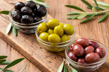 Mix of marinated olives in glass bowls on wooden cutting board. Green black and red kalamata olives with leaves. Mediterranean appetizer and healthy snack.