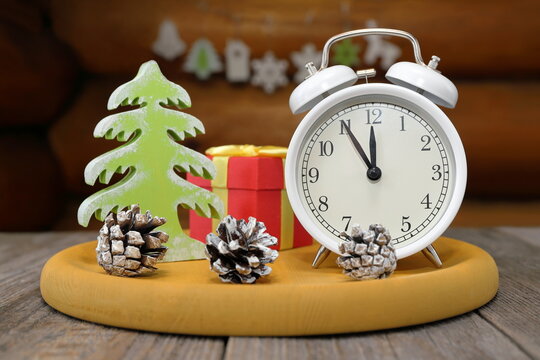 On a yellow tray, against log wall decorated with garland of plywood Christmas ornaments, stands white retro alarm clock that shows 5 minutes to 12, red gift box, wooden Christmas tree and pine cones.