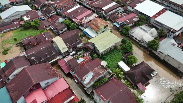 Aerial views captured by drones show residential areas submerged in floodwaters, densely packed houses with rusty, colourful roofs, muddy roads, and lush green trees.