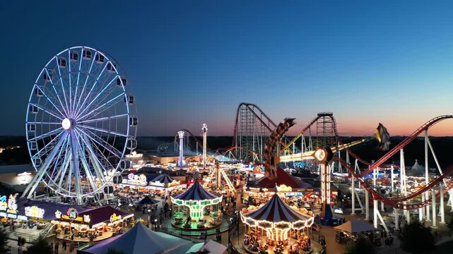 Illuminated amusement park at dusk with ferris wheel and roller coaster. Evening entertainment theme park with bright carnival lights.