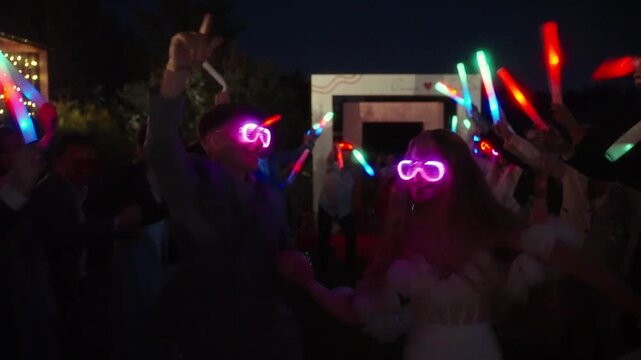 On a wedding party newlywed couple in glowing glasses walks through a crowd of guests holding colorful LED sticks at night. Diverse group of friends and relatives. Translation: Alex and Stacy