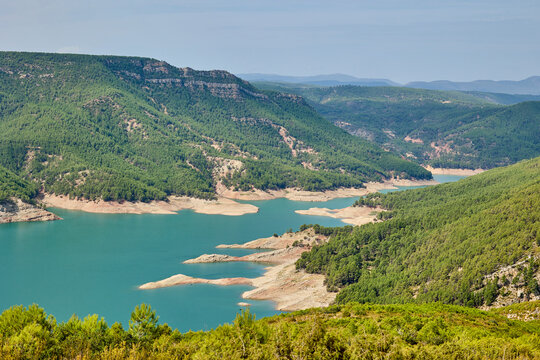 Wide panoramic view of a turquoise mountain lake surrounded by green forests and hills in Spain under a partly cloudy sky. Peaceful natural landscape and wilderness scenery. - Powered by Adobe