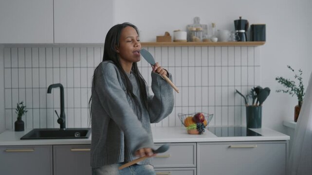 Funny african woman moving by music at home and dancing in kitchen, portrait. Cute female person singing song at kitchenware, enjoying good mood in holidays, stay positive and have fun in weekend