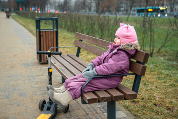 A little child girl in a hat and warm clothes sat down to relax on a bench, next to a scooter on a rainy cold day