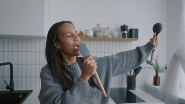 Pretty young woman enjoying music and singing son at home kitchen, good mood. Positive black lady having fun alone at home in weekend, be happy and feel joy, funny female person with kitchenware