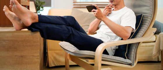 Relaxed young asian man using a smartphone while sitting comfortably in an armchair at living room