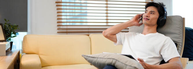 Young Asian man wearing headphones relaxing in cozy living room, smiling while choosing music on tablet