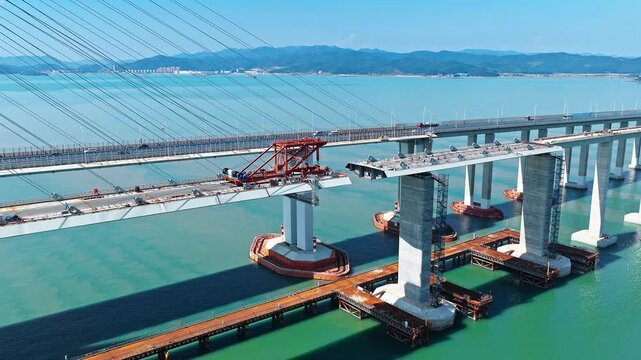 Aerial shot of massive sea-crossing bridge under construction with engineering machinery and traffic on existing highway 