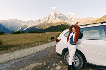 Fototapeta premium Woman by car in mountain landscape, smiling with arms raised beside white SUV on a gravel road, travel adventure in an open meadow with scenic peaks, clear sky and relaxed vibe.
