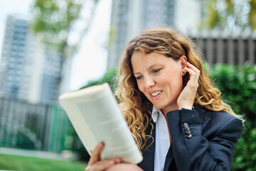 Portrait of a young businesswoman woman holding and reading a book sitting on a bench in park, surrounded by moder corporate office buildings