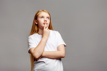 Thoughtful teenage girl with long golden blond hair looking up in studio, isolated on gray background, young female model in white clothing posing