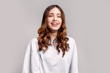 Beauty portrait of smiling teenage girl with glossy long curly hair, glowing skin and natural makeup, studio light on neutral background. Young female model with beautiful smile and teeth