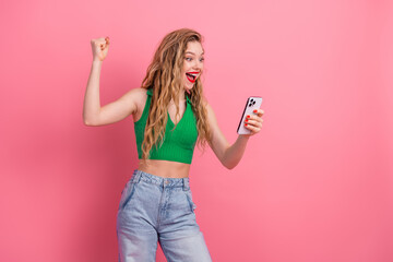 Excited young woman with blonde hair using a smartphone on a vibrant pink background celebrating...