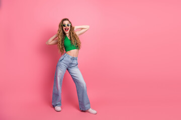 Cheerful young woman in stylish green top posing joyfully with hands raised on pink background