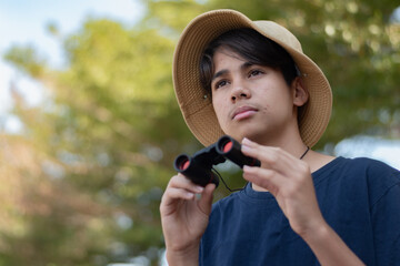 A close-up shot of a Thai teenage boy diligently wearing a cream bucket hat and holding binoculars around his neck, during field study in a park.