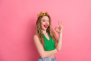 Charming young woman wearing a stylish crown making an OK hand gesture against a vibrant pink...