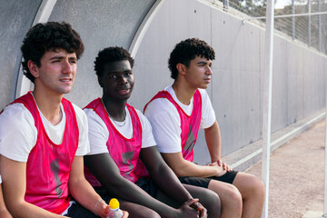 Three diverse male athletes in pink bibs resting on the bench, observing the game, anticipating their turn to play