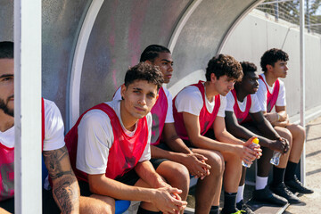 Young diverse male soccer team resting on the bench, hydrating and watching the game during a sunny outdoor match, focused and ready