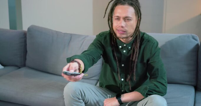 Man wanting show sitting on living room sofa pressing remote while sipping red juice and chuckling
