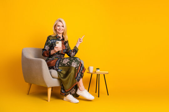 Elegant mature woman in a patchwork dress sits in a beige chair holding a phone against a bright yellow background