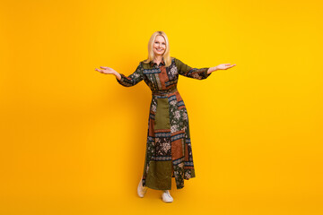 Happy mature woman in a patchwork dress stands with arms open against a yellow background