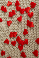 Small red satin hearts are spread across a background of textured beige knitted fabric. The hearts are randomly scattered, creating a Valentine's Day themed aesthetic.