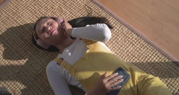 Mid adult Asian woman on rug placing speaker adjusting headphones feeling rhythm when music plays