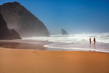 Two women walk through powerful ocean waves beside a dramatic cliff on a windy beach, capturing adventure, freedom, and wild coastal nature.