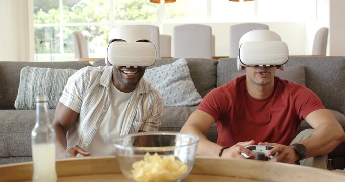 Diverse men donning VR headsets holding controllers on sofa at home laughing and playing VR game