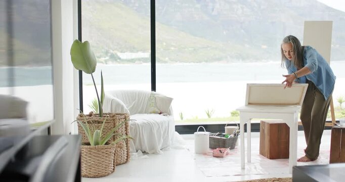 Senior woman opening wooden easel box, adjusting blank canvas at table by lake window, copy space
