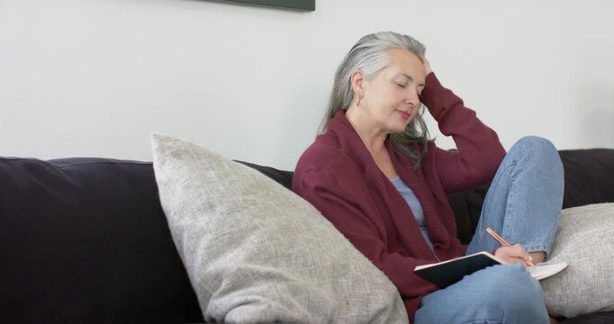 After settling on couch Senior woman reflectively journaling in notebook with pencil at home
