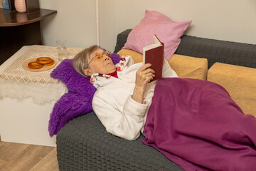 Elderly woman with eyeglasses relaxing on a couch, covered with a blanket, reading a book. Concept of comfortable retirement and leisure.