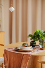 Table setting with rustic runner, white plate, green foliage, and hanging decor. Simple home interior.