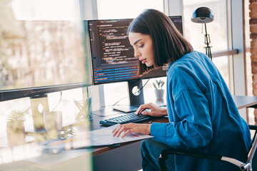 Female programmer works in a bright office coding at a dual monitor setup while planning projects